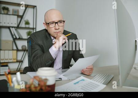 Mature businessman in suit sitting at the table with computer monitor and working with documents Stock Photo