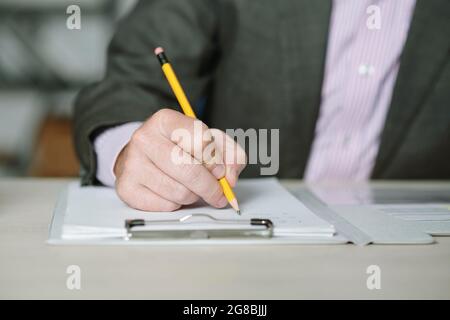 Close-up of businessman sitting at the table and making notes in document with pencil Stock Photo