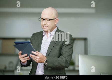 Mature bald businessman in suit holding digital tablet in his hands and working online at office Stock Photo