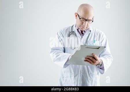 Mature doctor in white coat making notes in medicl card standing against the white background Stock Photo
