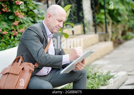Mature businessman making notes in documents while sitting on the bench outdoors Stock Photo