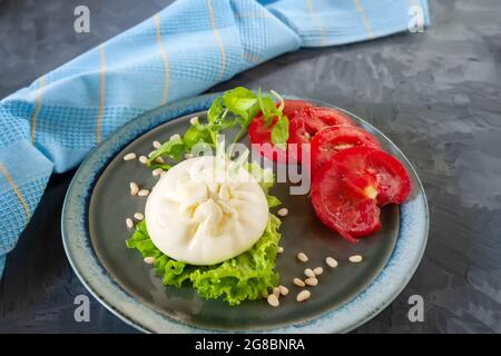 Fresh soft cream burrata cheese with tomatoes and green salad on a beautiful plate. There is a plate of cheese and a blue napkin on the table. Stock Photo