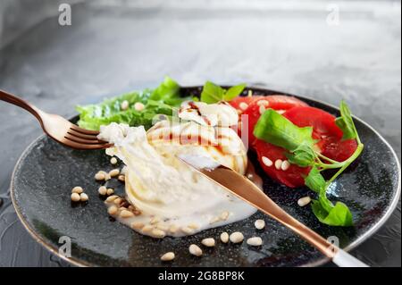 Fresh soft cream cheese burrata with tomatoes and green salad on a beautiful plate. On the table there is a plate with cut cheese and cutlery, a fork Stock Photo