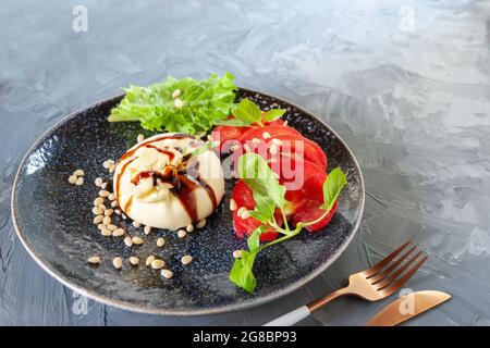 Fresh soft cream cheese burrata with tomatoes and green salad on a beautiful plate. On the table is a plate of cheese and cutlery, a fork and a knife. Stock Photo