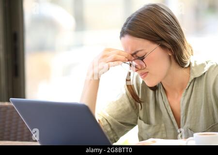 Tired woman wearing eyeglasses suffering eyestrain using laptop sitting in a bar Stock Photo