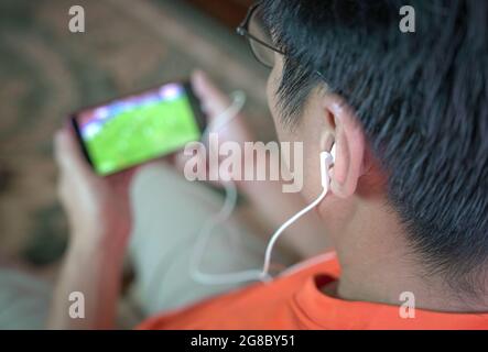 Man watching football and sport stream on mobile phone. Selected focus on ear phones. Stock Photo