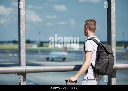 Traveler watching airplanes at airport. Rear view of passenger with backpack looking through window of terminal building. Stock Photo