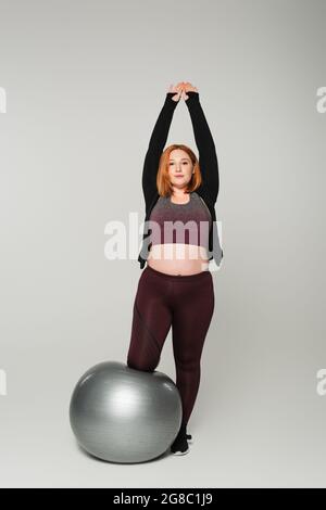 Redhead plus size sportswoman stretching near fitness ball on grey background Stock Photo
