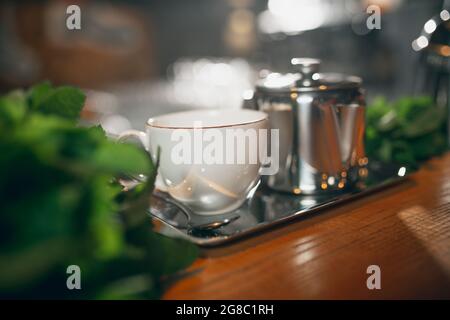 Cafe style. Image of white cup and milk jug standing in metal tray on wooden table behind bunches of mint, greens. Stock Photo
