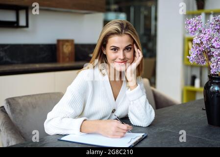 A young woman is working in her kitchen and is writing something in a notebook. Businesswoman works remotely from home. Stock Photo