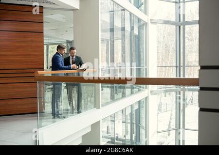 Two businessmen discussing document together while standing in the corridor of the modern office building Stock Photo