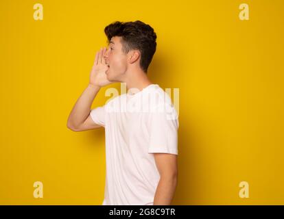 Announcement. Handsome young man holding hands near opened mouth. Isolated on yellow. Stock Photo