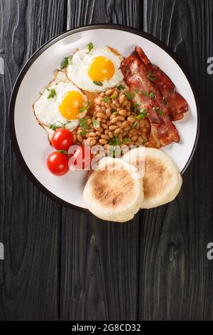 Hot hearty breakfast of fried eggs, beans, bacon, tomatoes and muffins close-up in a plate on the table. vertical top view from above Stock Photo