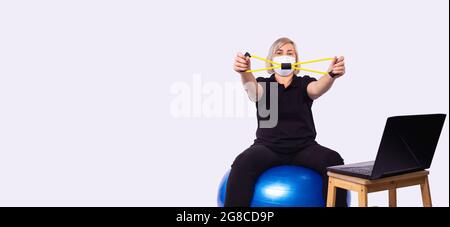 Woman in black sportswear and face medical mask having online training while watching the tablet and doing fitness exercises. Stock Photo