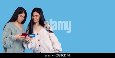 Banner, long format. Twins girls in knitted sweaters are holding cellphone and enter data from the card. Stock Photo