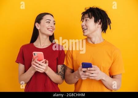 Young multiracial couple smiling and using mobile phones isolated over yellow wall Stock Photo