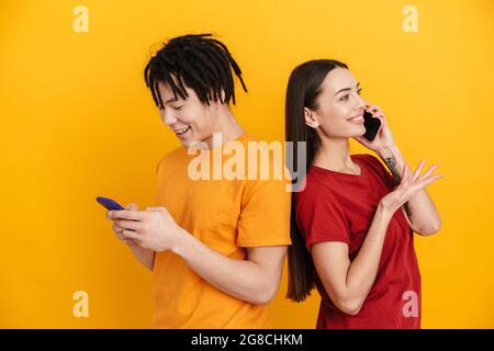 Young multiracial couple smiling while using and talking on cellphones isolated over yellow wall Stock Photo