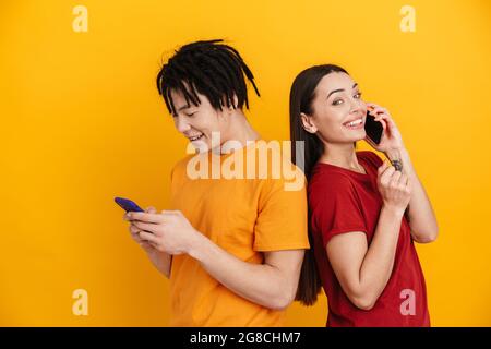 Young multiracial couple smiling while using and talking on cellphones isolated over yellow wall Stock Photo
