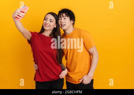 Smiling multiracial couple hugging while taking selfie on cellphone isolated over yellow wall Stock Photo