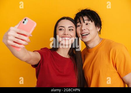 Happy young multiethnic couple standing over yellow background, taking a selfie Stock Photo