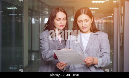 Young women entrepreneurs in grey jackets discuss new project looking at sheets of papers in building hall with light reflections, sunlight Stock Photo