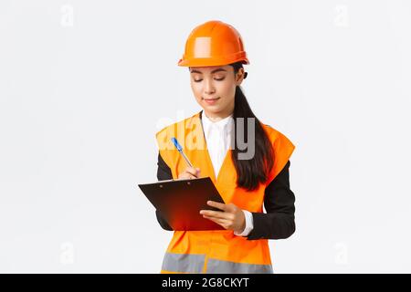 Pleased asian female construction engineer, architect taking notes in clipboard, writing down something during inspection at building area, wearing Stock Photo