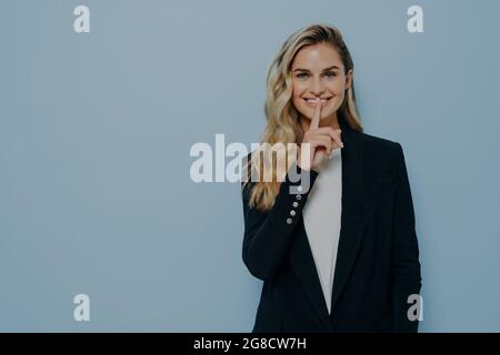 Cheerful beautiful young woman keeps finger on lips and asking to keep something in secret or be silent Stock Photo