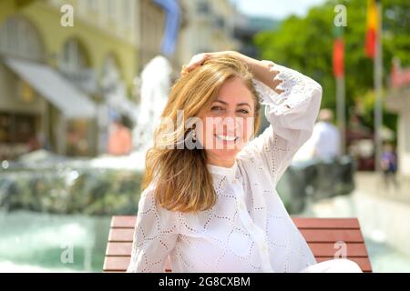 Happy attractive woman relaxing on a chair in the hot sunshine holding her hand to her long hair as she looks to the camera Stock Photo