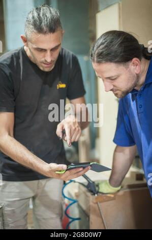 Carpenters resting in workshop and scrolling on smartphone. Handsome handicraftsman texting message and tapping on phone Stock Photo