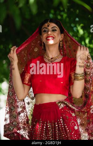 cheerful indian bride in red sari and traditional headscarf with ornament Stock Photo