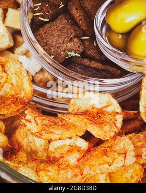 Selected focus, close up, vertical, food composition with salty snacks: rye crackers, wheat crackers, potato chips with spices and green olives in gla Stock Photo