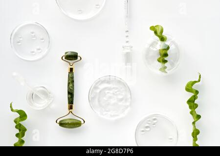 Self made moisturizer and green jade face roller. Exotic fern leaves and water drops on white background. Facial massage, handmade cosmetics. Stock Photo