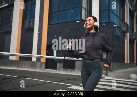 Smiling joyous sportswoman in wireless headphones and activewear jogging ahead Stock Photo