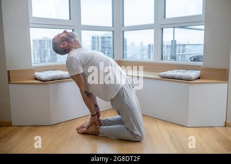 Gray-haired man in a white tshirt doing yoga and looking involved Stock Photo