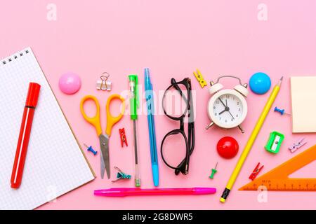 School notebook and office supplies on a pink background. Back to school abstract background. White alarm clock and multi-colored pens and black glasses. Stock Photo