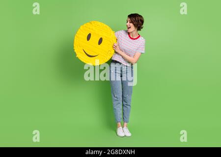 Full length body size view of pretty cheerful girl holding in hands emoji sticker opinion isolated over green pastel color background Stock Photo