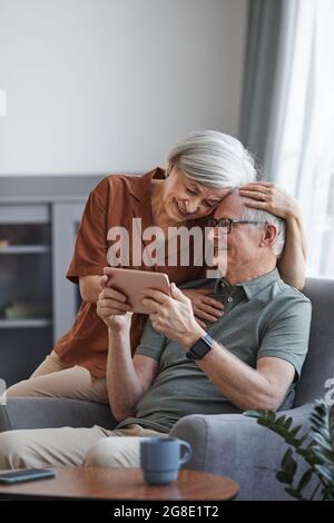 Vertical portrait of happy senior couple using digital tablet together at home and embracing Stock Photo