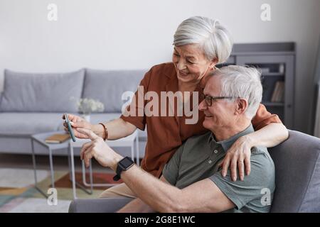 Side view portrait of happy senior couple using smartphone together at home and embracing, copy space Stock Photo