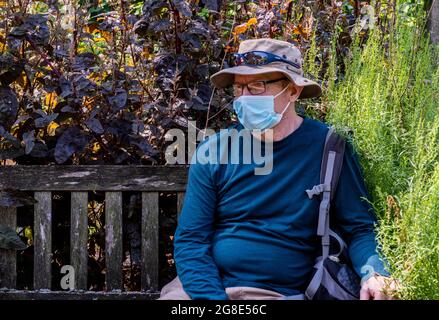 A man wearing face mask, glasses and a straw hat sitting on a bench in the Chelsea Physic Garden in London. Stock Photo