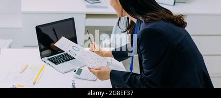 Cheerful Asian woman in jacket surfing laptop and holding paper while standing by desk and whiteboard with graphs in modern office Stock Photo