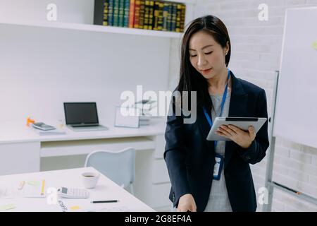 Intent Asian woman in jacket surfing tablet and smile while standing by desk in modern office Stock Photo