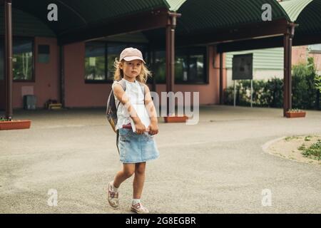 The cute girl, elementary school student, walking to school with bag behind back and book. Students are ready for the new year. Back to school. Stock Photo