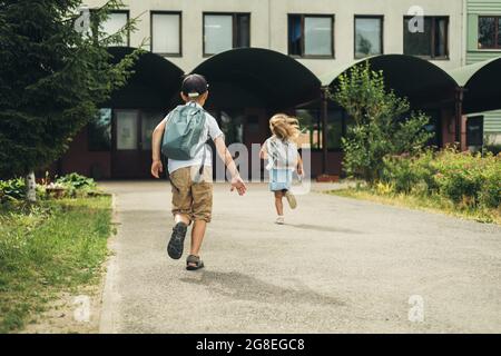 Two caucasian children, boy and girl, running to school with bags behind their backs. Students are ready for the new year. Back to school. Stock Photo