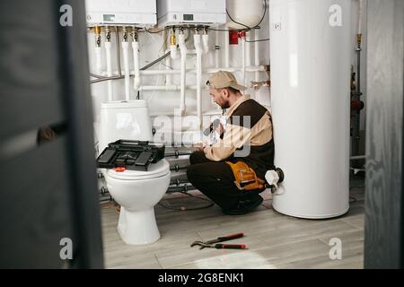 Young repairman in uniform examining part of pipe in toilet Stock Photo