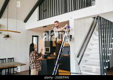 Young worker on stepladder talking to female client while fixing alarm system Stock Photo