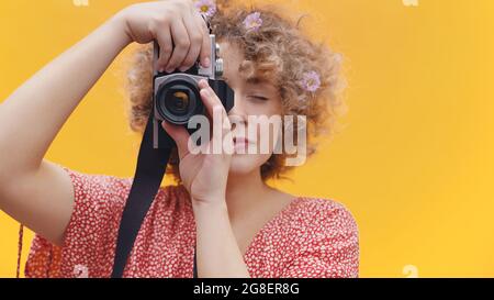 Attractive girl holding a vintage camera taking a picture. Girl in a joyful mood over bright yellow background studio. Dressed in a casual pink shirt wearing flowers in her hair. Photography concept. Stock Photo