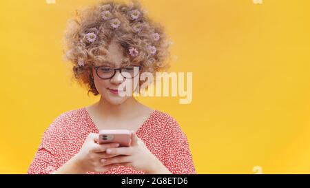 Attractive girl wearing nerdy spectacles holding a mobile phone in her hand. Typing a text on her cellphone and smiling. Concept of online text messages and social media. Bright yellow background studio.  Stock Photo