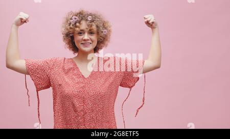 Attractive girl showing her biceps. Making a fist with both hands. Smiling and feeling proud. Concept of power and strength. Dressed in a casual pink shirt. Isolated in a pink background studio. Stock Photo