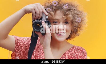 Attractive girl holding a vintage camera taking a picture. Girl in a joyful mood over bright yellow background studio. Dressed in a casual pink shirt wearing flowers in her hair.  Stock Photo