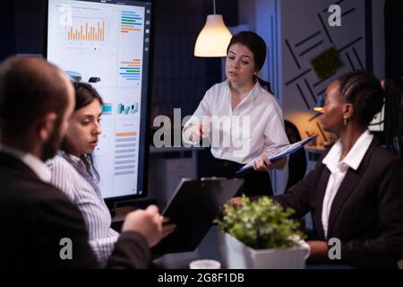 Diverse multi ethnic teamwork brainstorming company ideas analyzing financial graphs presentation. Businesspeople overworking in meeting room late at night discussing solution Stock Photo